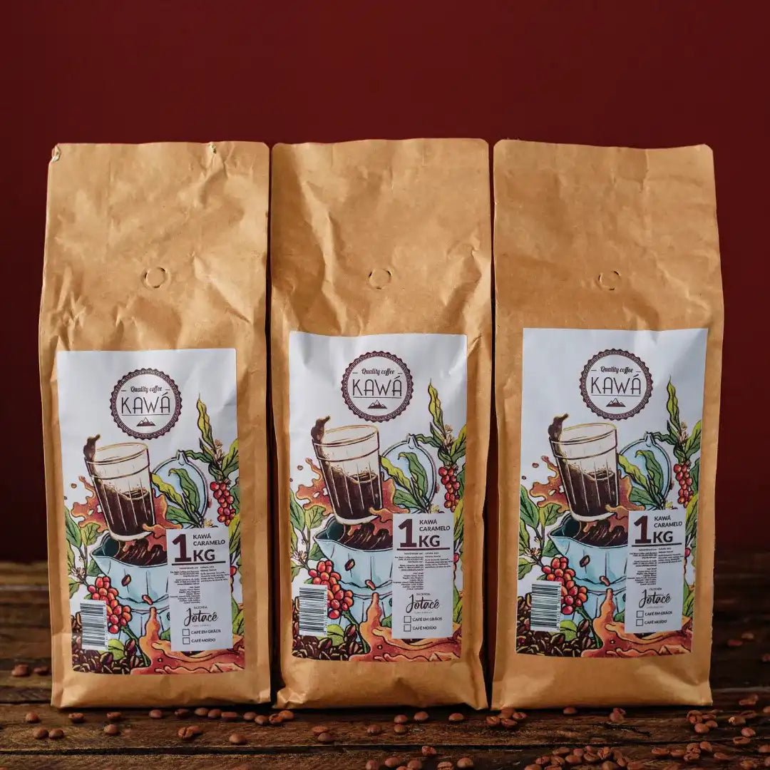3 pacotes de 1 Kg em pé do produto Kawá Caramelo dispostos lado a lado sobre mesa de madeira com grãos soltos