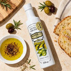 Garrafa de Azeite de Oliva Extra Virgem Prosperato Premium 250 mL deitada sobre bancada clara, com tigelinha de azeite temperado com especiarias, fatias de pão, azeitonas e ramos de alecrim ao redor.
