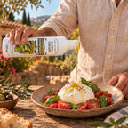 Mão regando um prato de burrata com tomates e manjericão usando o Azeite Extra Virgem Prosperato Premium 250 mL, numa mesa ao ar livre com paisagem de oliveiras ao fundo.