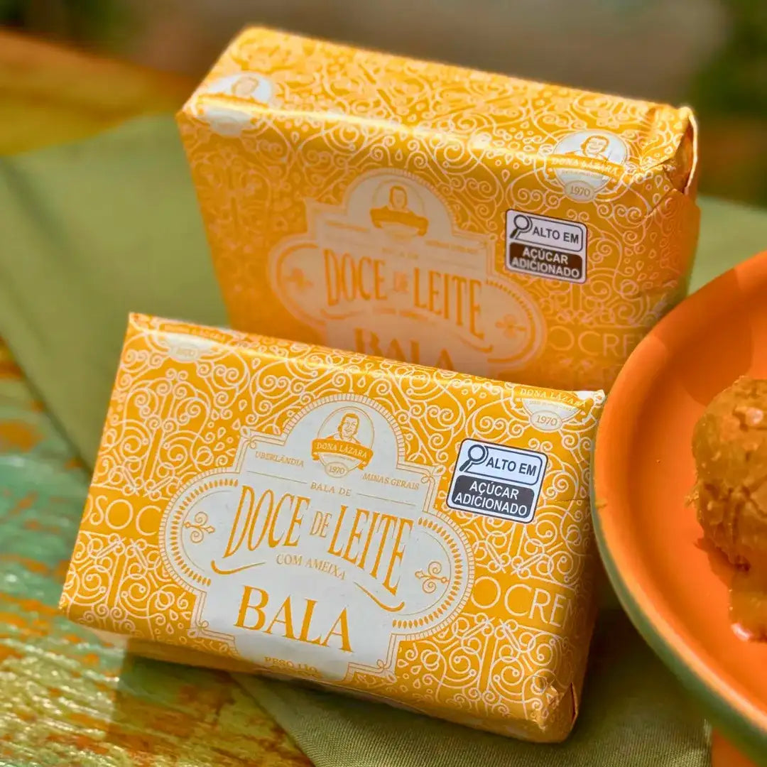 Duas embalagens da Bala de Doce de Leite com Ameixa Puxa-Puxa Dona Lázara em embalagem amarela sobre mesa com toalha verde, ao lado de prato com pedaço do doce
