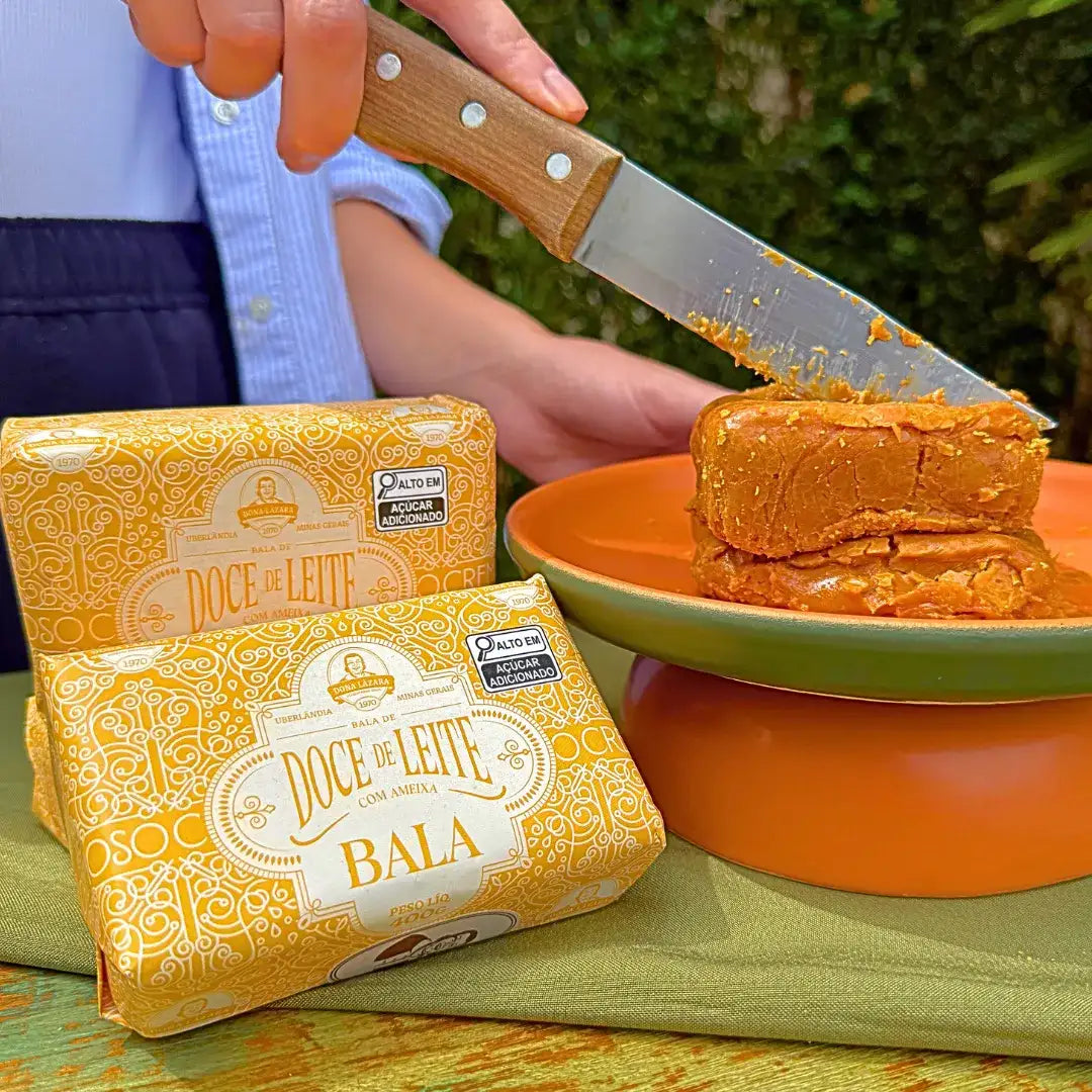 Embalagens da bala puxa-puxa de ameixa Dona Lázara ao lado de pedaço cortado com faca de madeira sobre prato laranja, revelando a textura cremosa do doce artesanal