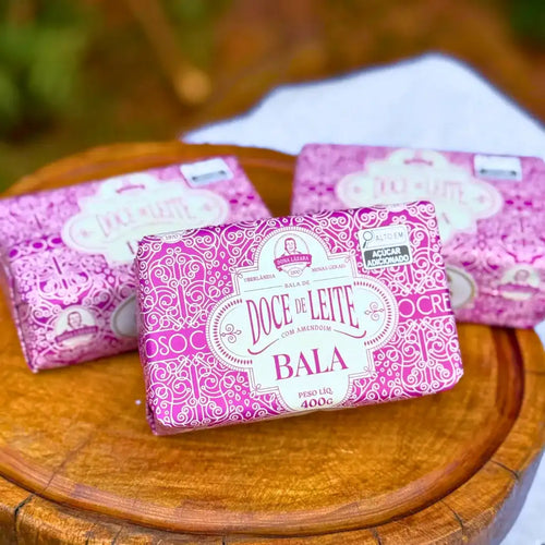 Três embalagens rosa da Bala de Doce de Leite com Amendoim Puxa-Puxa Dona Lázara 400g dispostas sobre tábua de madeira redonda, com detalhes da identidade visual da marca