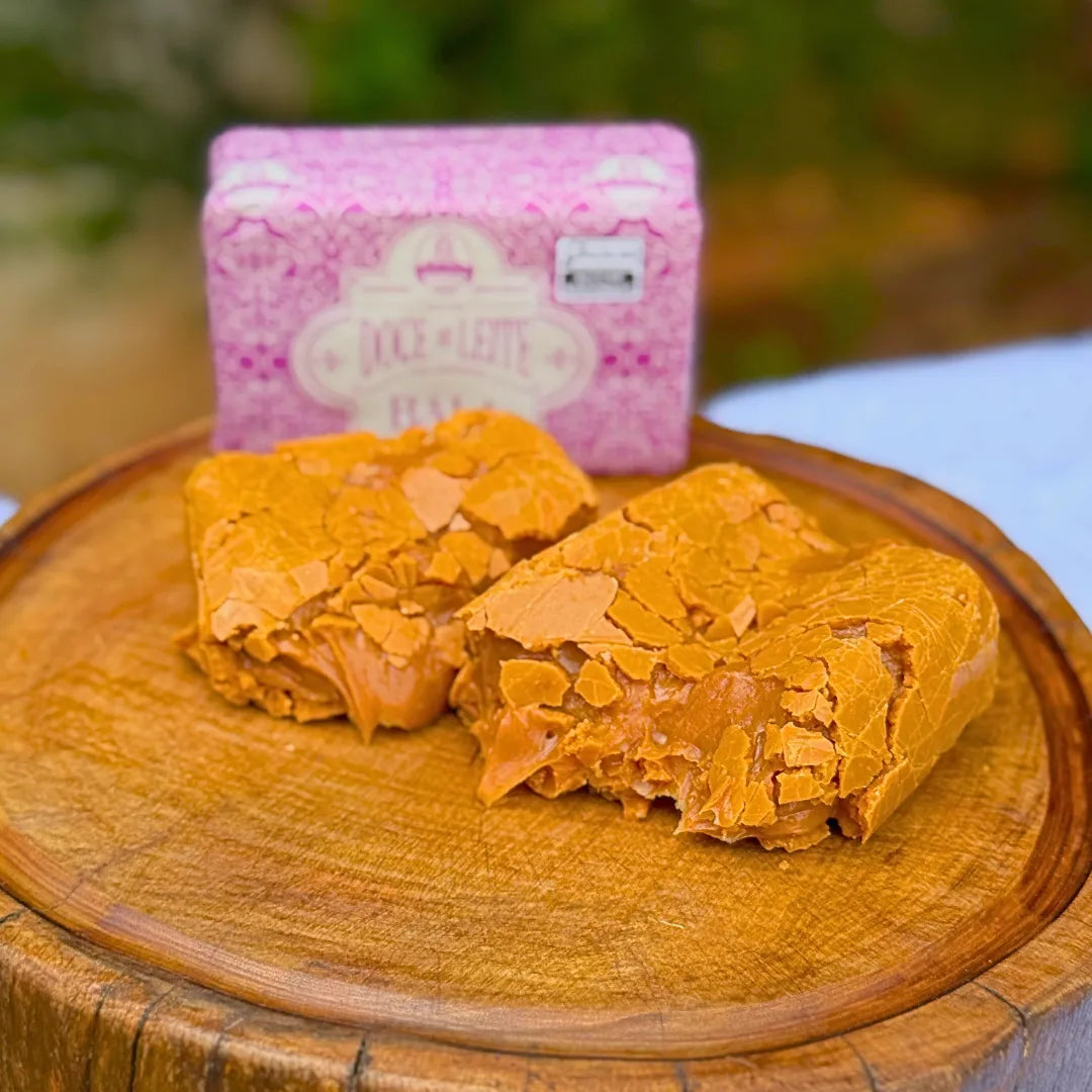 Pedaços de bala puxa-puxa de amendoim artesanal partida sobre tábua de madeira, com textura crocante e cremosa visível, e embalagem rosa Dona Lázara ao fundo
