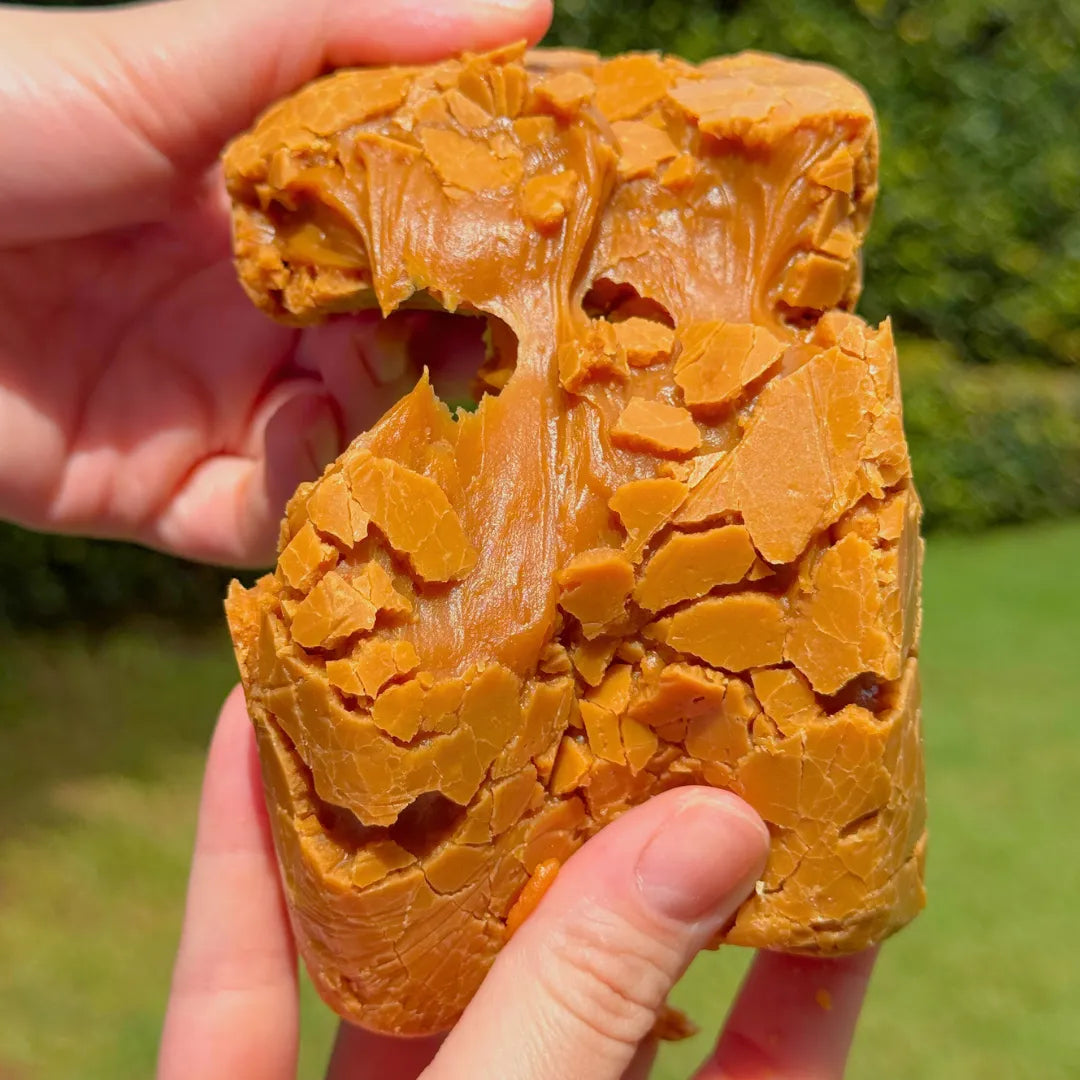 Close de mãos abrindo bala puxa-puxa de amendoim artesanal, exibindo camadas crocantes do doce de leite com amendoim da Fazenda Jotacê sobre fundo de gramado verde