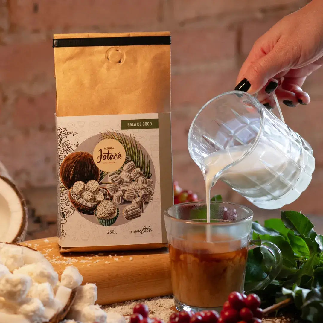Leite sendo adicionado ao café Bala de Coco da Fazenda Jotacê, com ambientação de cocos e grãos de café sobre tábua de madeira.