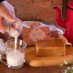  Pessoa servindo leite na caneca, com pedaço de bala de doce de leite puxa-puxa Dona Lázara sobre a tábua, embalagens ao fundo e bule vermelho, em clima aconchegante de fazenda.