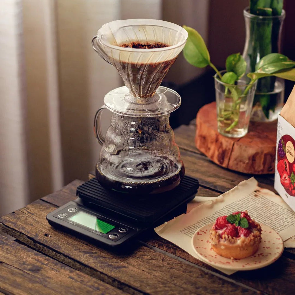 Balança digital black em funcionamento extraindo café com uma jarra de vidro e filtro V60 ao lado de bolo em mesa de madeira