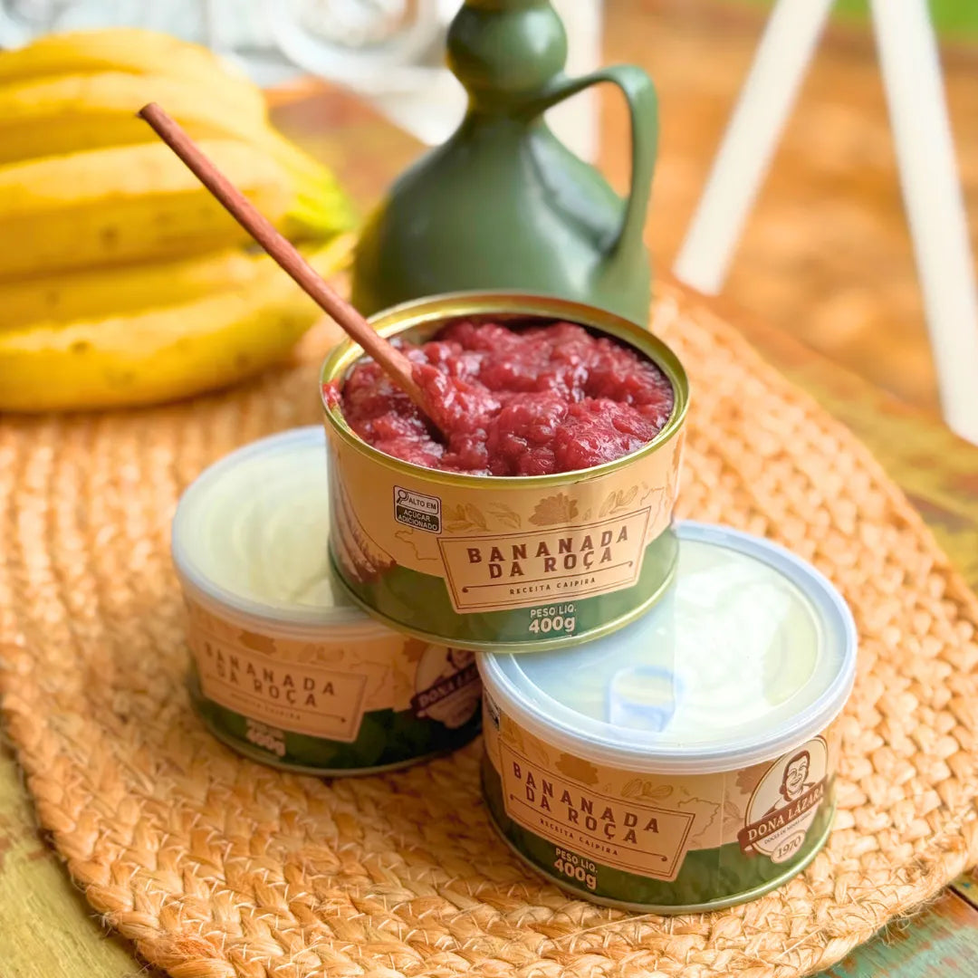 Três latas de Bananada da Roça Dona Lázara 400g sobre sousplat de palha trançada, com lata aberta mostrando a bananada cremosa e colher de pau, cacho de bananas ao fundo