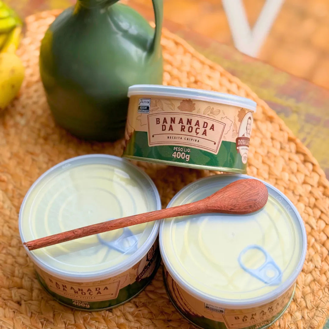 Latas de Bananada da Roça Dona Lázara fechadas mostrando o lacre e colher de pau de madeira, com moringa verde decorativa e bananas ao fundo em cenário rústico