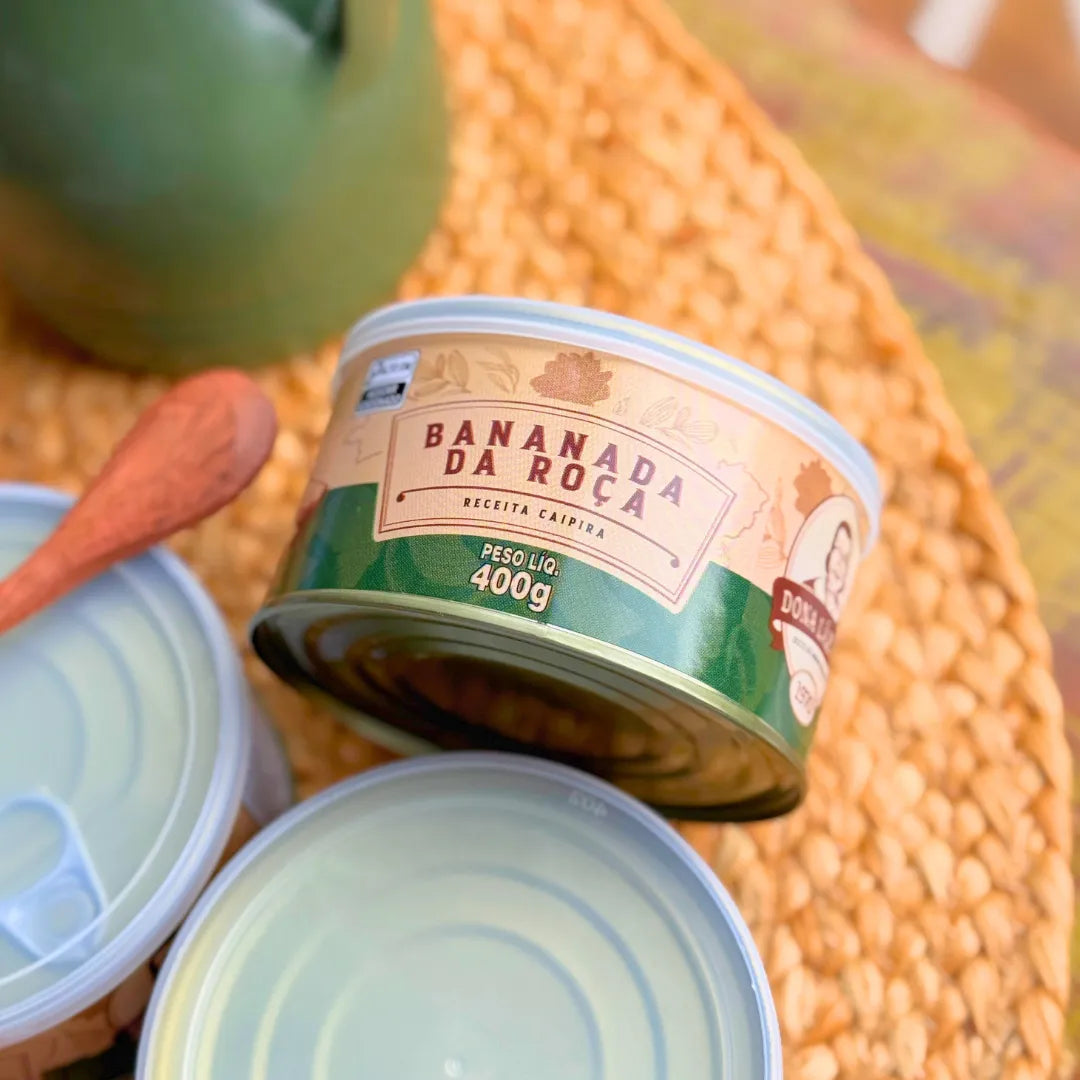 Lata fechada de Bananada da Roça Dona Lázara 400g em ângulo superior sobre sousplat de palha trançada, mostrando rótulo com logotipo Dona Lázara e informações do produto