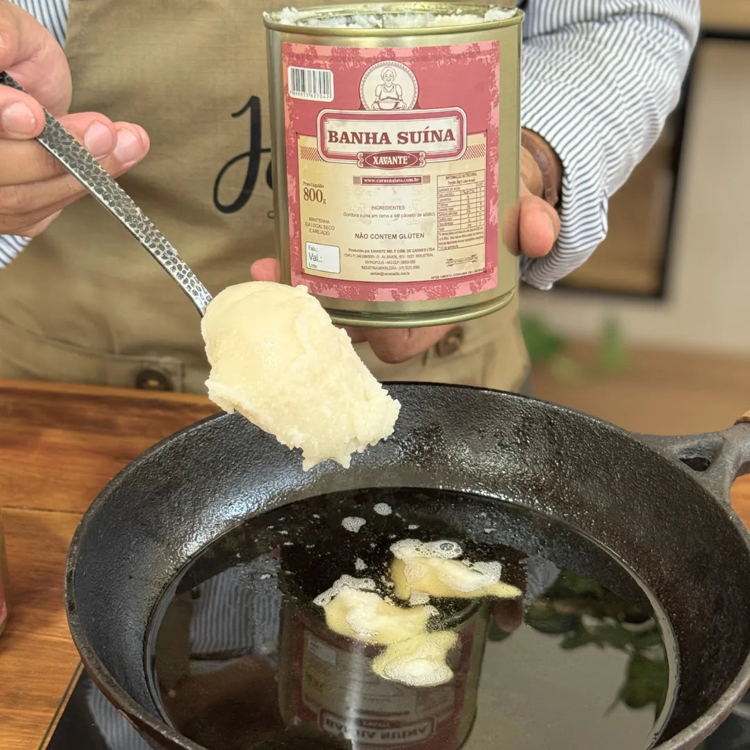 anha suína Xavante 800g sendo colocada em frigideira de ferro com colher, com a lata da banha de porco segurada ao fundo na cozinha.