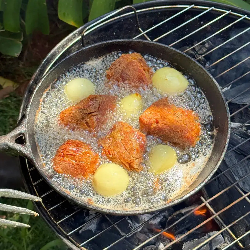Carne suína sendo frita na banha de porco Xavante em panela de ferro sobre fogareiro a carvão, com cebolas douradas borbulhando na gordura.