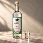 Garrafa de cachaça branca ao lado de um copinho servido, sobre bancada de mármore ao sol