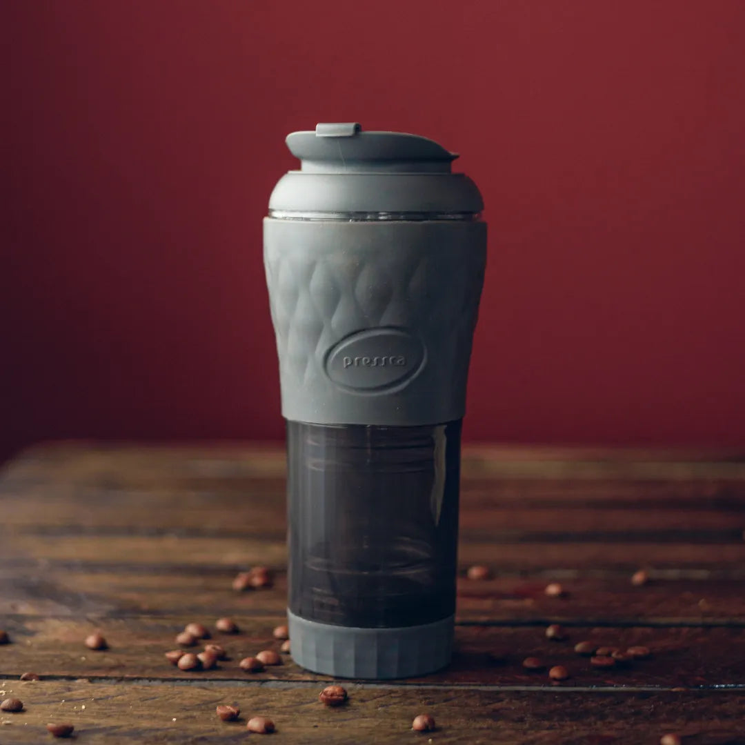 Cafeteira Portátil Pressca cinza com grãos de café ao redor sobre mesa de madeira.