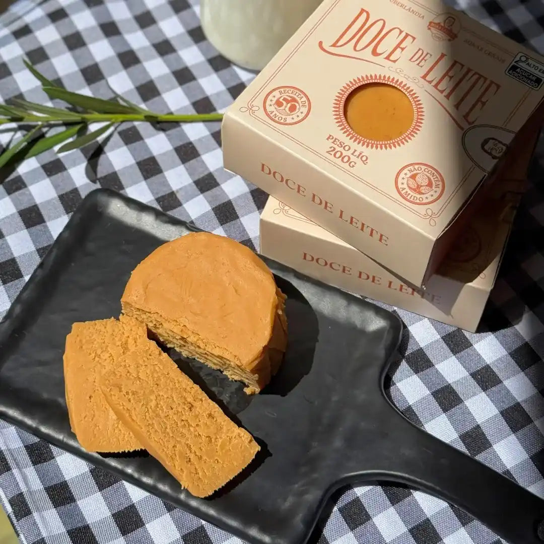 Doce de leite de corte fatiado sobre tábua preta, com caixas do produto ao fundo.