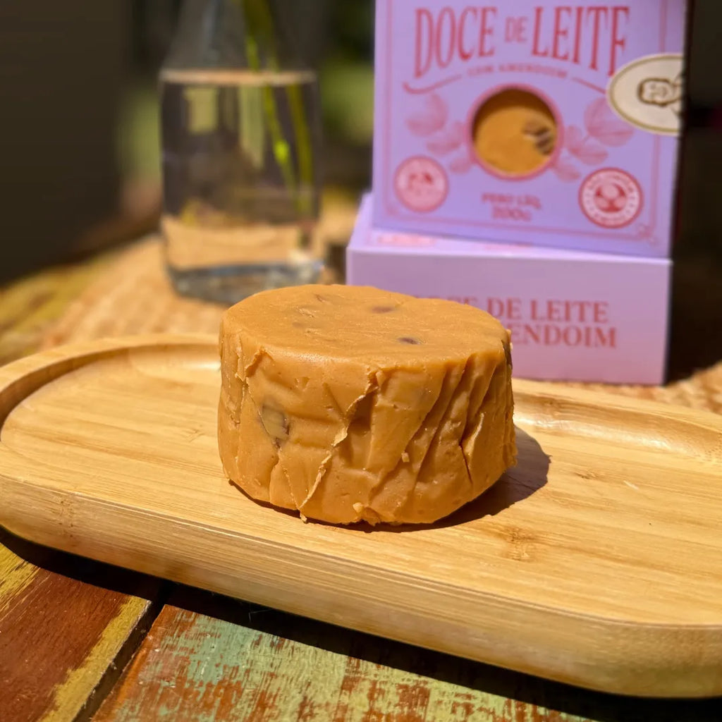 Peça inteira de doce de leite com amendoim sobre bandeja de madeira, com a caixa gaveta lilás desfocada ao fundo.