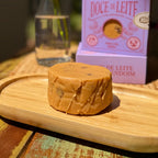 Peça inteira de doce de leite com amendoim sobre bandeja de madeira, com a caixa gaveta lilás desfocada ao fundo.