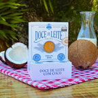 Duas caixas de Doce de Leite com Coco Dona Lázara com coco fresco e leite ao redor, sobre toalha xadrez, em composição que destaca o visual artesanal e o sabor caseiro.