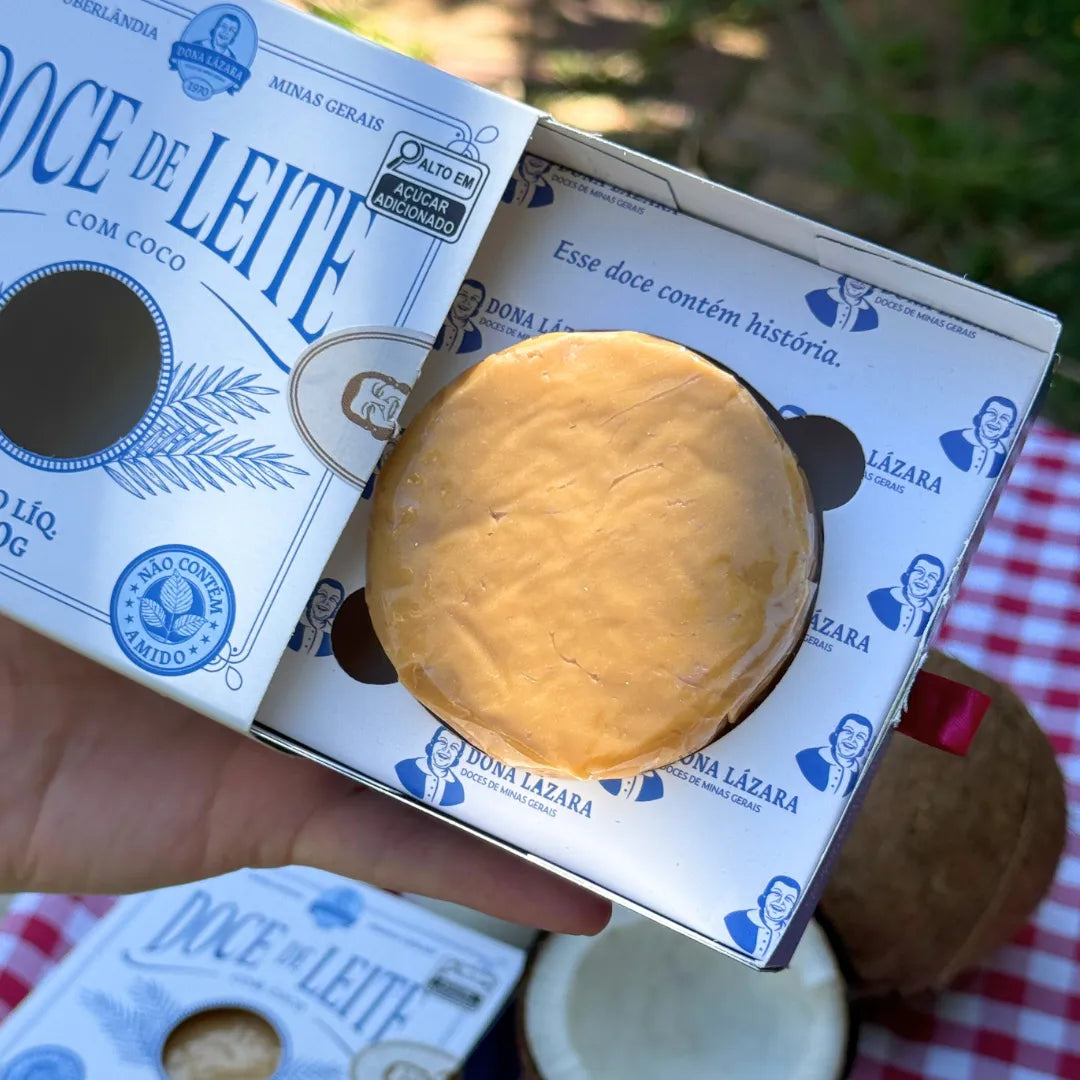 Close do Doce de Leite com Coco Dona Lázara sendo aberto, revelando o doce redondo e dourado dentro da caixa artesanal com a frase “Esse doce contém história”.