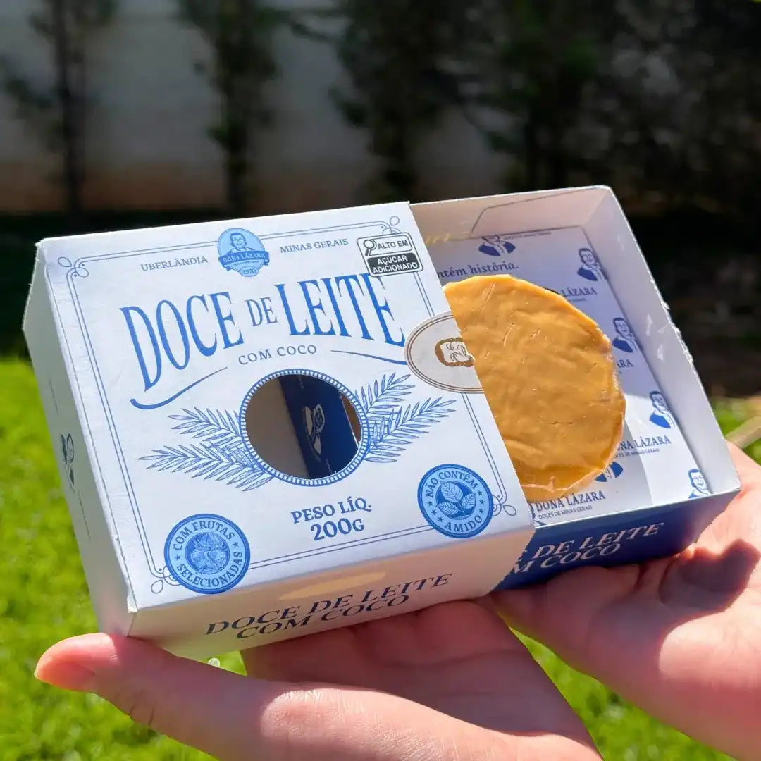 Caixa de Doce de Leite com Coco Dona Lázara sendo segurada nas mãos, sob a luz do sol, destacando o design clássico e o doce artesanal embalado.