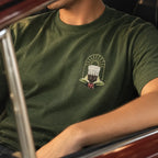 Close da camiseta verde militar vestida por um homem, mostrando a estampa pequena no peito com o copo de café coado, arco de sol e ramo de café.