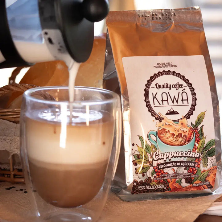 Leite sendo servido em uma xícara de vidro transparente com camadas visíveis, ao lado da embalagem do Cappuccino Kawá Zero Açúcar, com pães ao fundo.