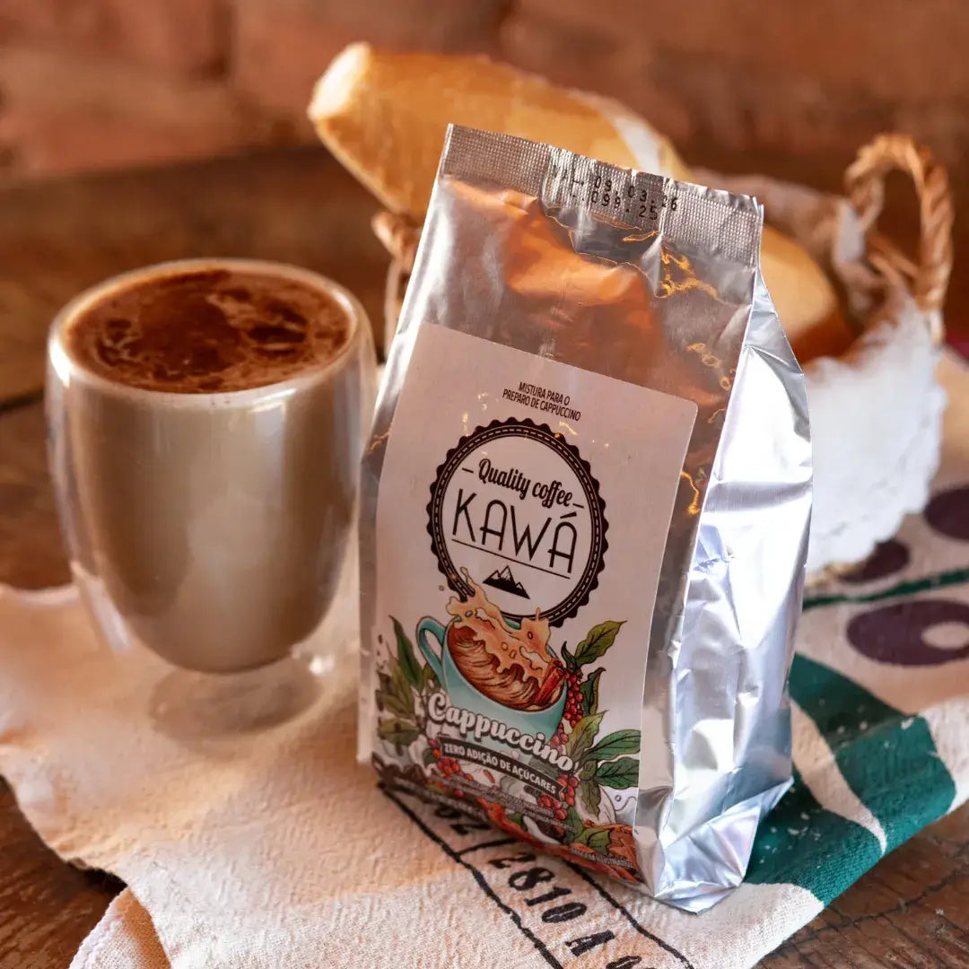 Xícara de cappuccino com canela por cima ao lado da embalagem do Cappuccino Kawá Zero Açúcar, sobre pano rústico com estampa, em mesa de madeira.