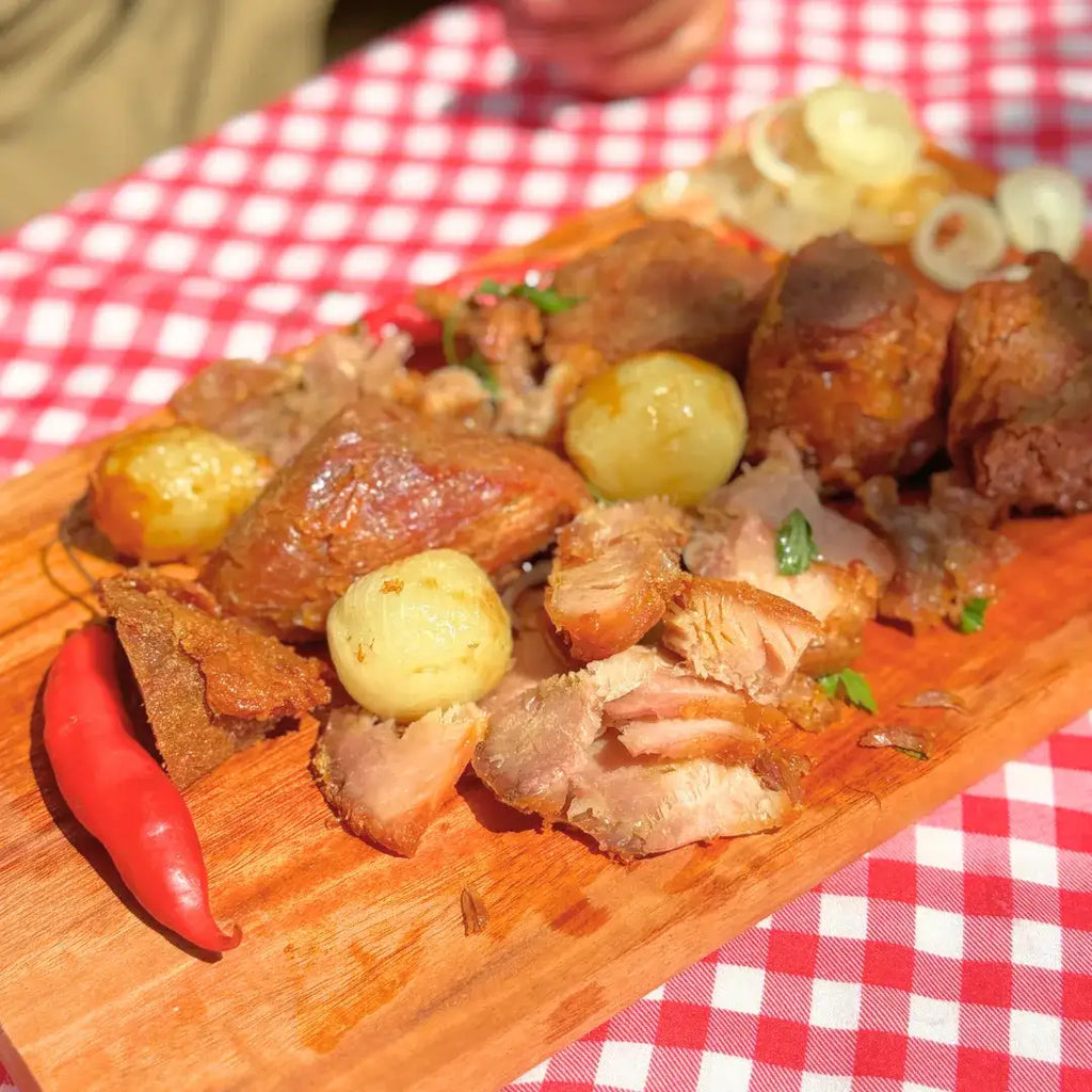 Pernil desfiado da carne na lata Xavante sobre tábua de madeira, acompanhado de batatas bolinhas, anéis de cebola e pimenta vermelha.