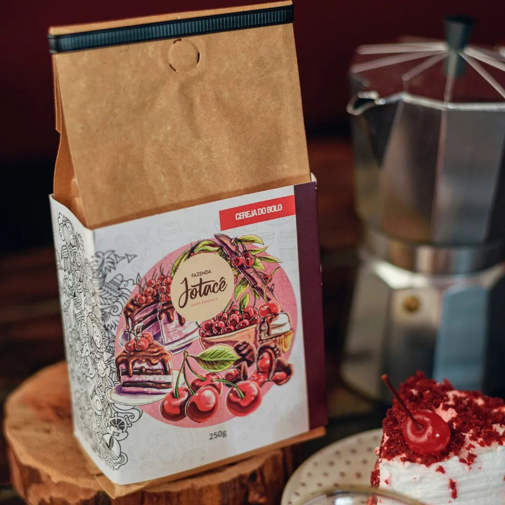 Pacote do Café Cereja do Bolo da Fazenda Jotacê ao lado de um bolo e com cafeteira italiana desfocada sobre mesa de madeira