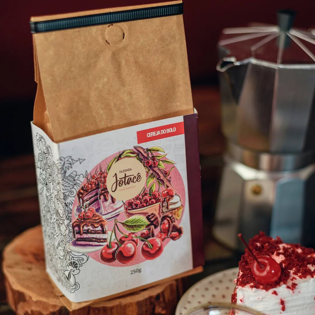 Pacote do Café Cereja do Bolo da Fazenda Jotacê ao lado de um bolo e com cafeteira italiana desfocada sobre mesa de madeira