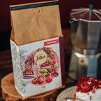 Pacote do Café Cereja do Bolo da Fazenda Jotacê ao lado de um bolo e com cafeteira italiana desfocada sobre mesa de madeira