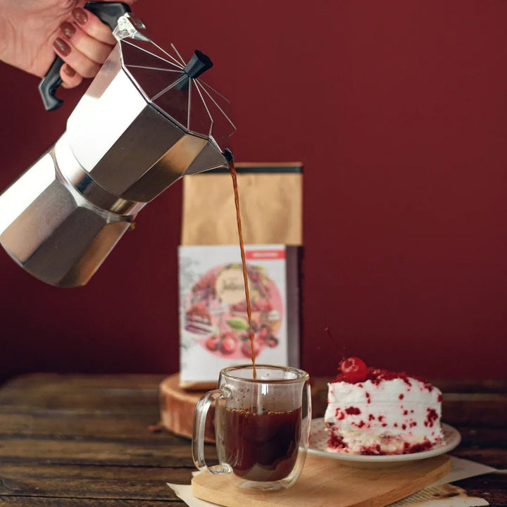 Cafeteira italiana despejando café em xícara ao lado de bolo e do Pacote do Café Cereja do Bolo sobre mesa de madeira