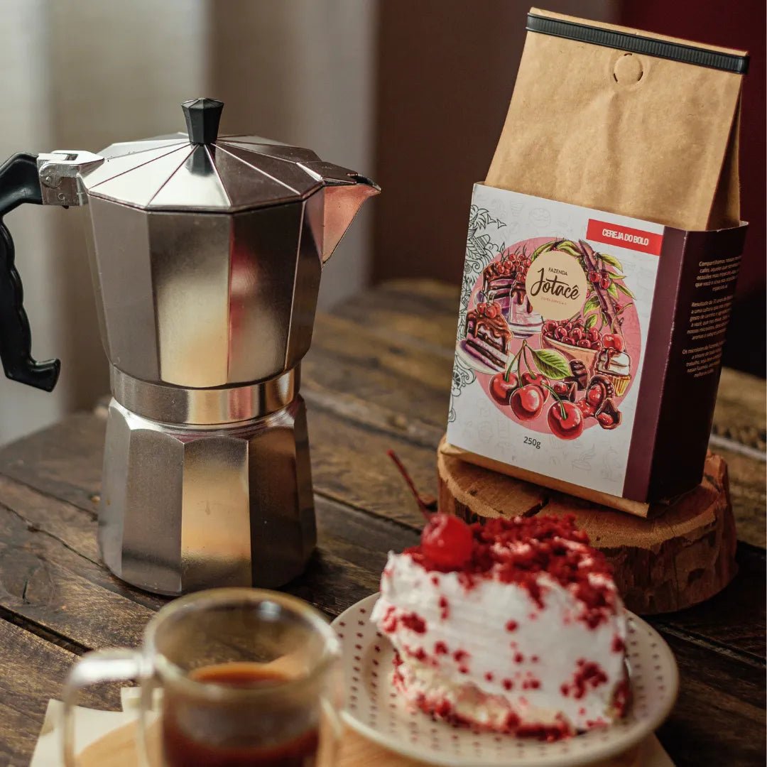Pacote do Café Cereja do Bolo da Fazenda Jotacê junto a uma cafeteira italiana com bolo e xícara sobre mesa de madeira