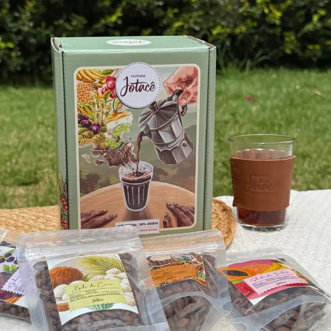 Caixa da Coleção Origens da Fazenda Jotacê ao ar livre, com copo americano de couro e pacotinhos de café especial sobre toalha clara.