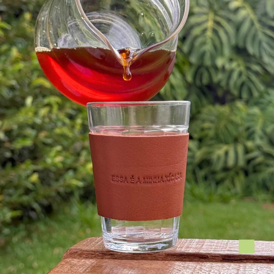 Copo americano com cinta de couro sendo preenchido com café fresco, com a frase gravada “Essa é a minha xícara” visível no couro.