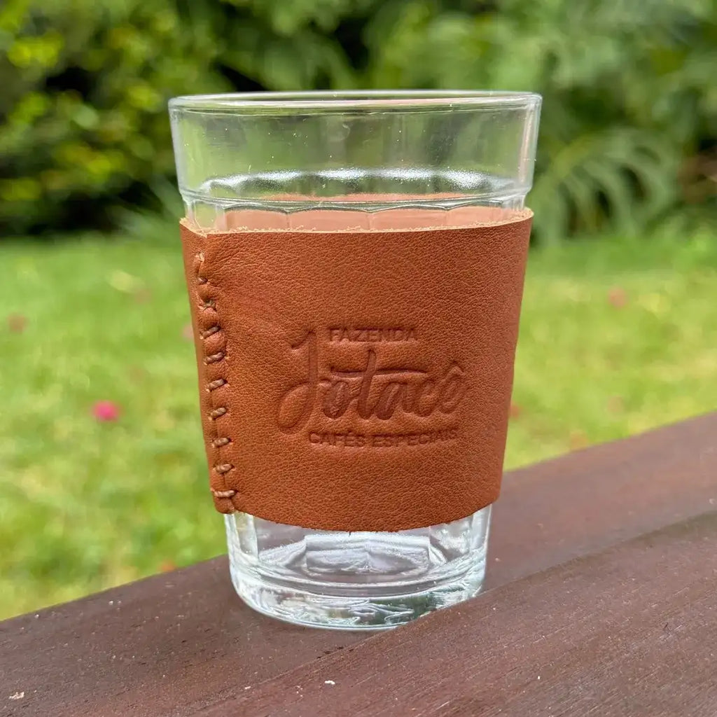 Copo americano com cinta de couro da Fazenda Jotacê em detalhe, sobre superfície de madeira com fundo verde de jardim.