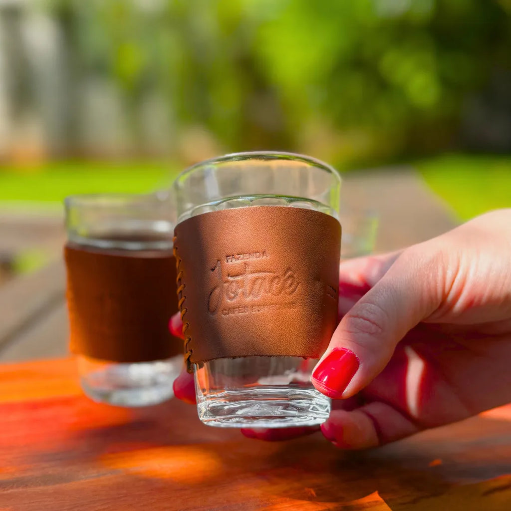 Mão segurando um copo com cinta de couro caramelo Fazenda Jotacê com café, ao lado de outro copo sobre a mesa de madeira, num brinde ao ar livre com verde ao fundo.
