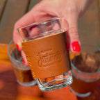 Mão com unhas vermelhas segurando o copo com cinta de couro Fazenda Jotacê, com a gravação "Fazenda Jotacê Cafés Especiais" em destaque e outros copos ao fundo.