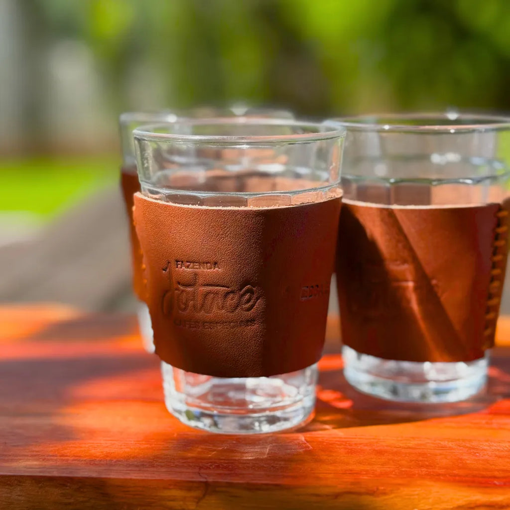 Dois copos de vidro com cinta de couro Fazenda Jotacê cheios de café sobre mesa de madeira ao ar livre, com a gravação na cinta visível e jardim desfocado ao fundo.