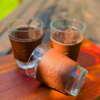 Três copos de vidro com cinta de couro caramelo Fazenda Jotacê sobre mesa de madeira ao sol, dois em pé com café e um deitado mostrando a gravação na cinta.