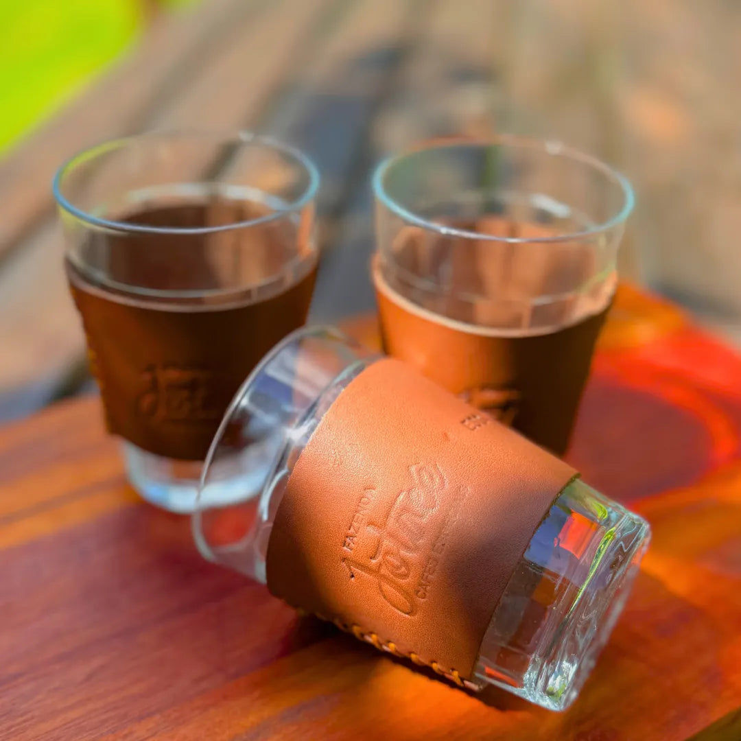 Três copos de vidro com cinta de couro caramelo Fazenda Jotacê sobre mesa de madeira ao sol, dois em pé com café e um deitado mostrando a gravação na cinta.