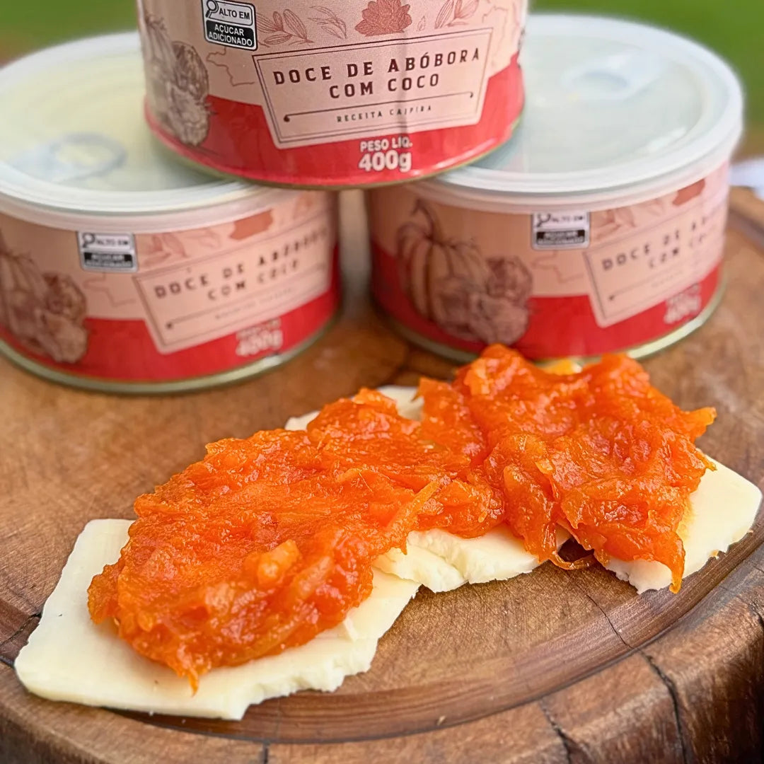 Doce de abóbora com coco artesanal servido sobre fatia de queijo minas em tábua de madeira, com três latas Dona Lázara 400g empilhadas ao fundo, combinação típica mineira