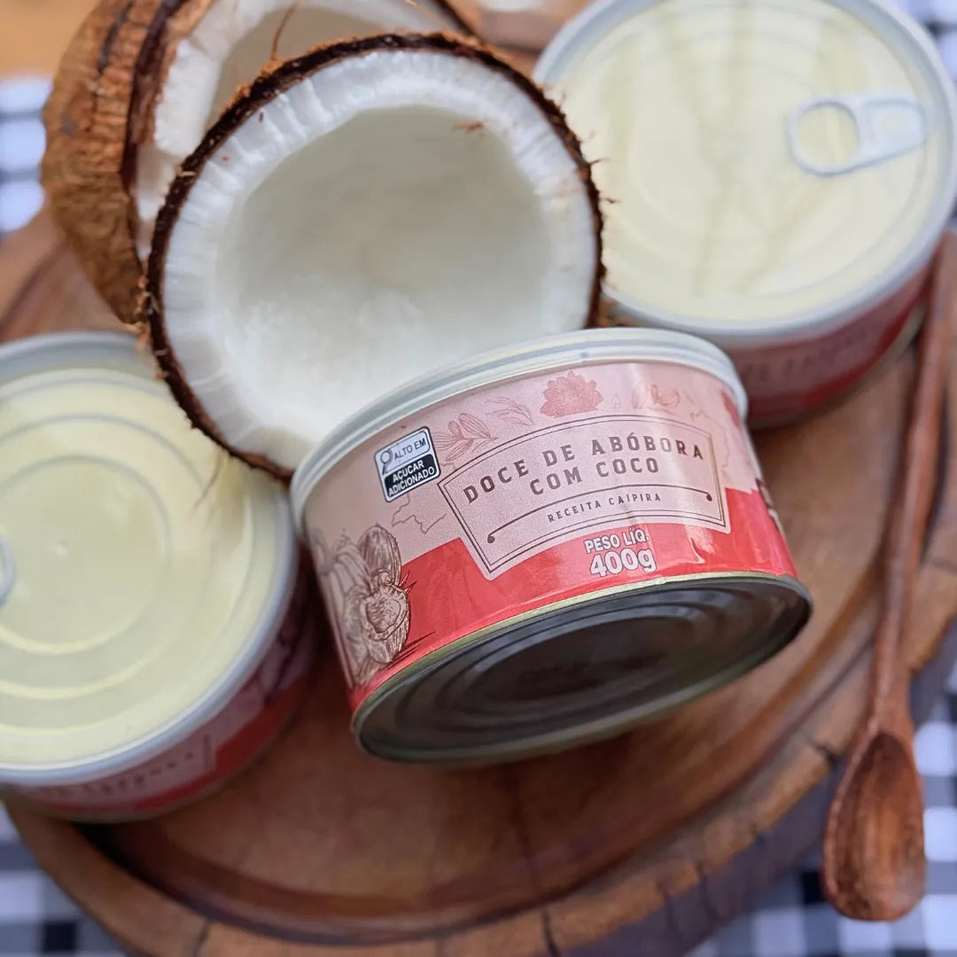 Latas de Doce de Abóbora com Coco Dona Lázara 400g vistas de cima sobre tábua de madeira, com coco partido ao meio ao lado e colher de pau, embalagem rosa com rótulo artesanal
