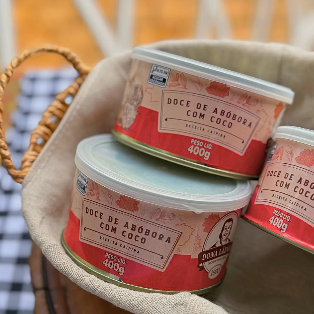 Três latas de Doce de Abóbora com Coco Dona Lázara 400g dentro de cesta de palha com tecido de linho, rótulo rosa com logotipo da marca e indicação de receita caipira