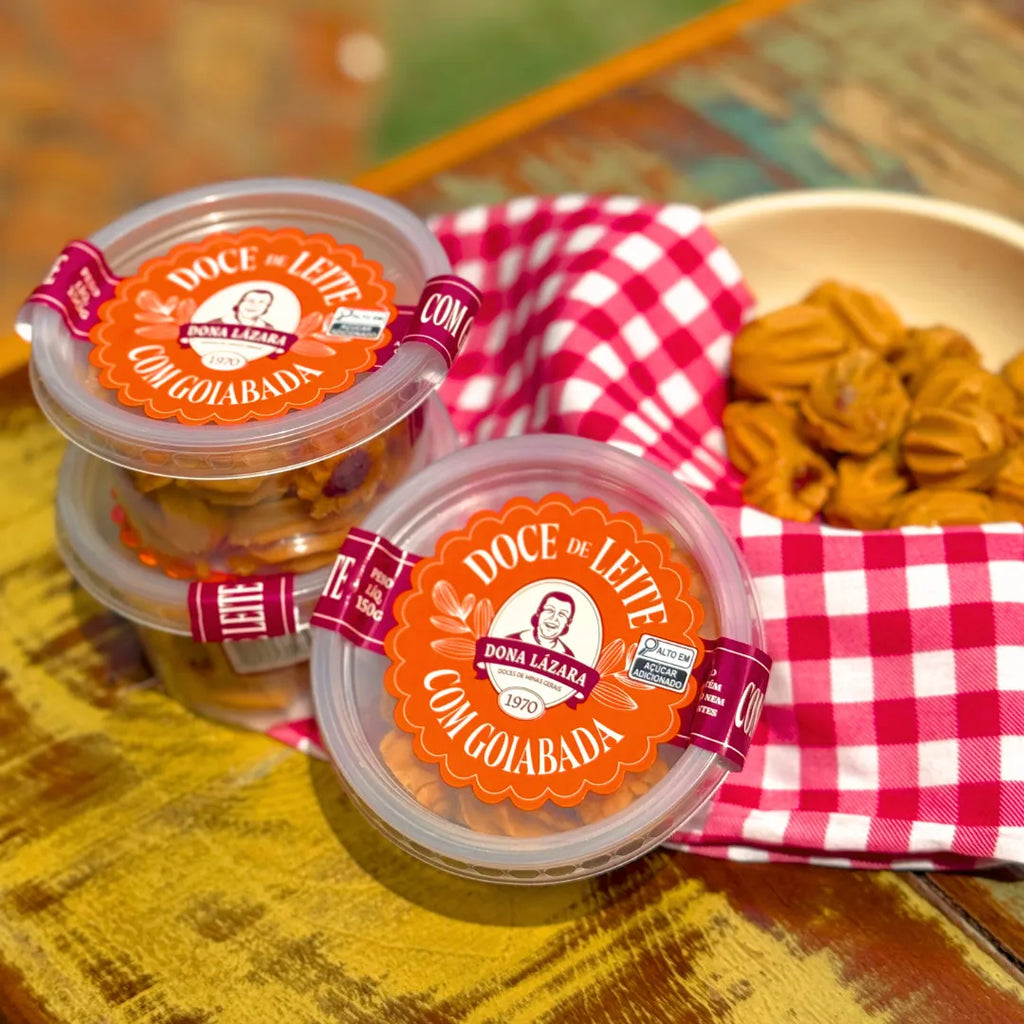 Dois potes de Doce de Leite com Goiabada Dona Lázara 150g sobre mesa rústica amarela, ao lado de tigela com docinhos moldados à mão e guardanapo xadrez vermelho