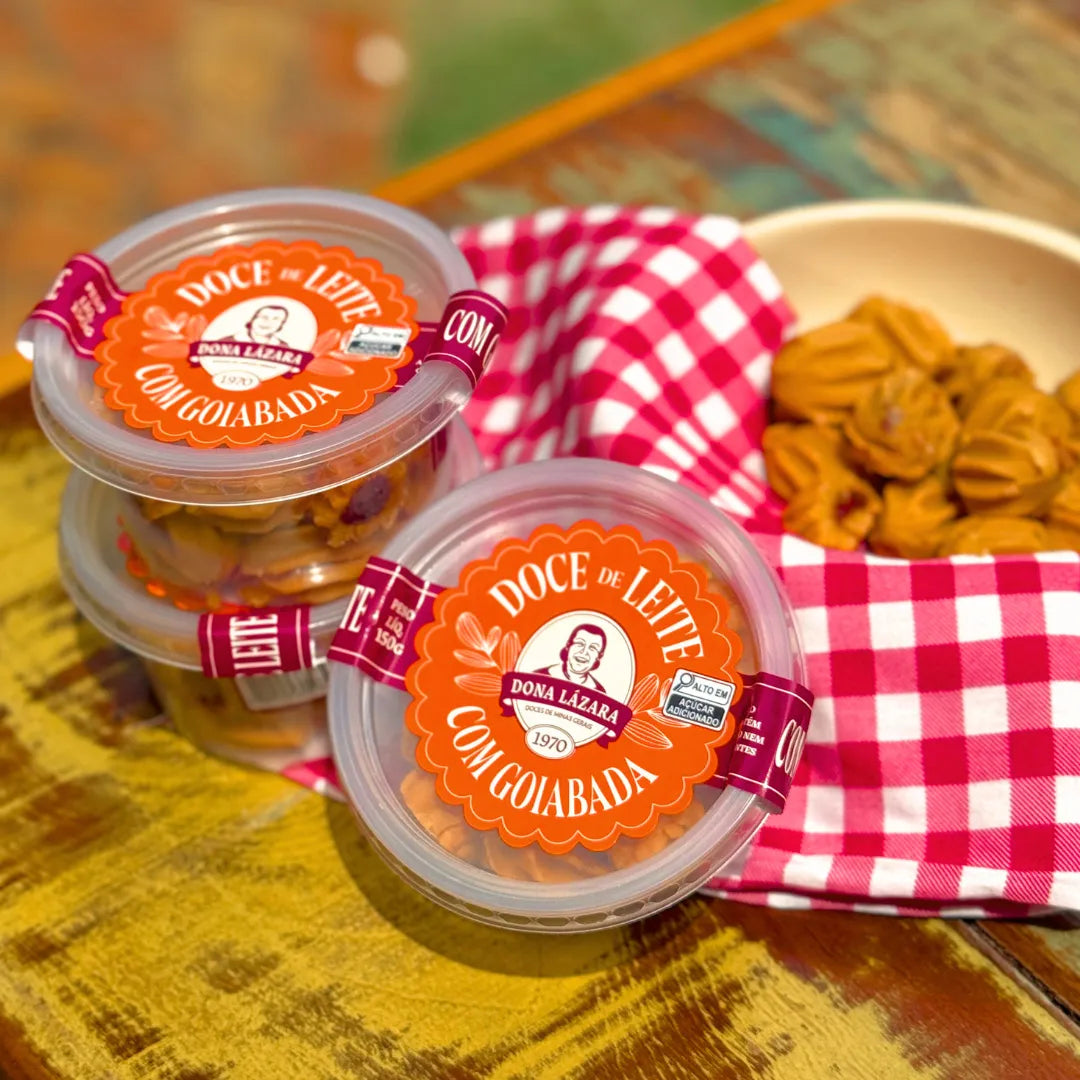 Dois potes de Doce de Leite com Goiabada Dona Lázara 150g sobre mesa rústica amarela, ao lado de tigela com docinhos moldados à mão e guardanapo xadrez vermelho