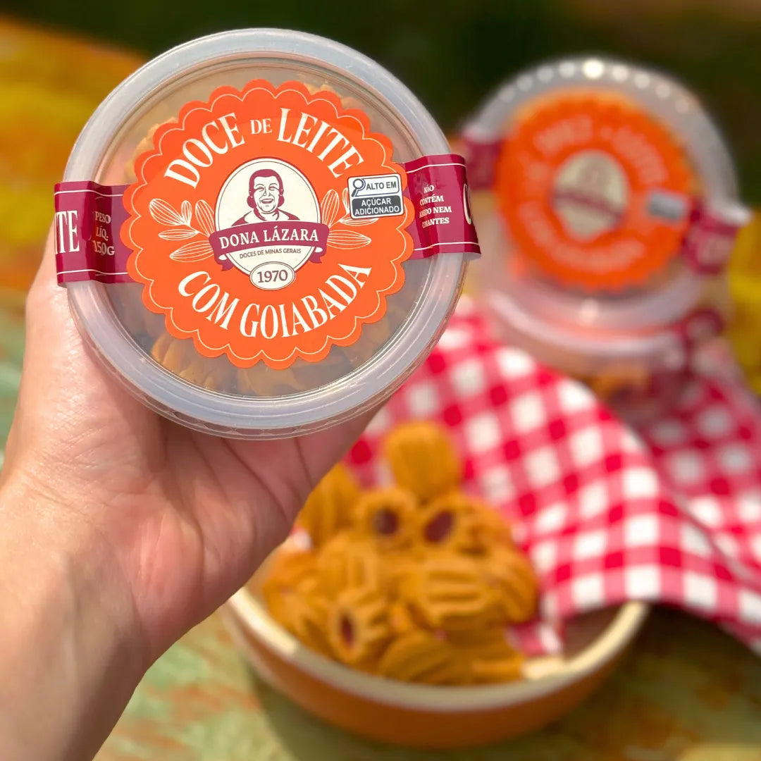 Mão segurando pote de Doce de Leite com Goiabada Dona Lázara 150g mostrando rótulo frontal com logotipo da marca desde 1970, docinhos artesanais desfocados ao fundo