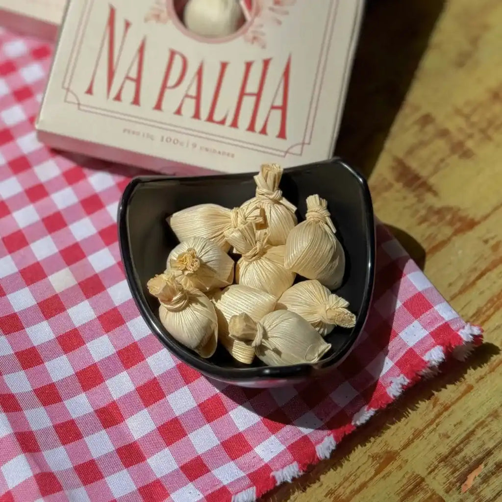 Tigela preta com várias unidades de Doce de Leite na Palha fechadas, posicionada sobre pano xadrez vermelho com a caixa do produto ao fundo.