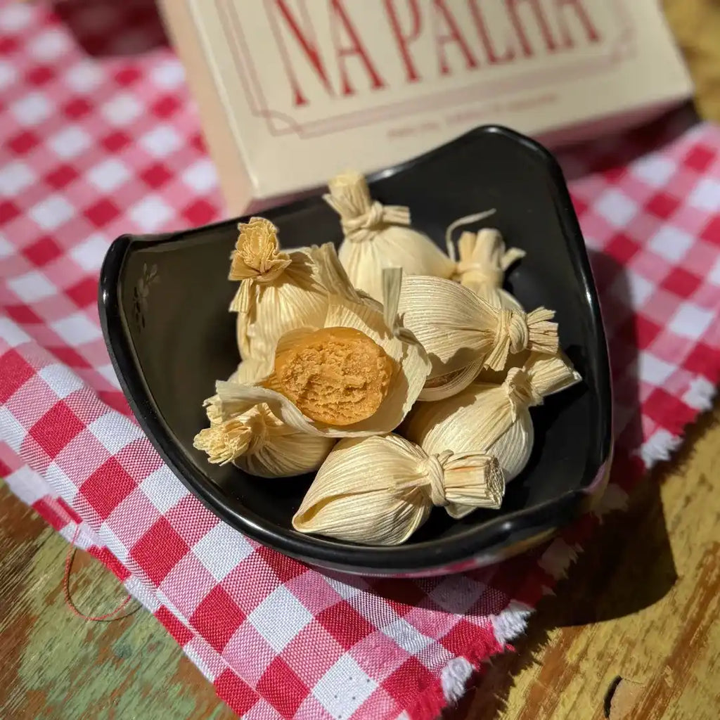Tigela preta com unidades de Doce de Leite na Palha abertas e fechadas, colocada sobre pano xadrez vermelho ao lado da caixa do produto.
