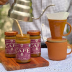 Potes de Doce de Leite com Café Dona Lázara 400g ao lado de um café sendo coado em coador de cerâmica, com xícara e bule